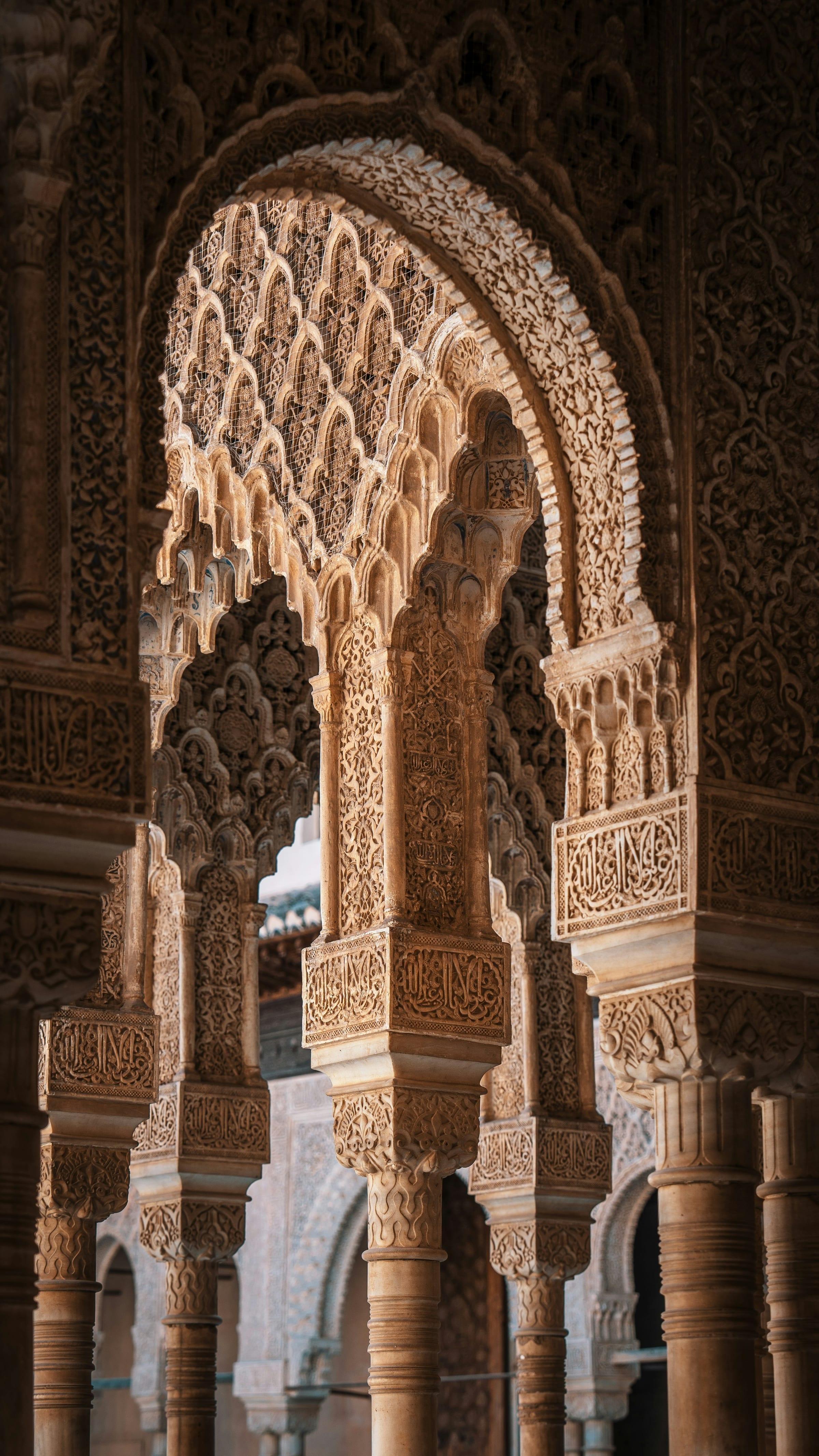 Intricately carved stucco arches and slender columns in the Alhambra, muqarnas honeycomb vaulting and bands of Arabic calligraphy layered above a horseshoe arch.